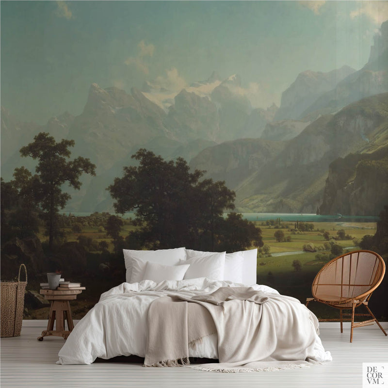 Alpine lake mountain wallpaper mural of misty Swiss peaks above a teal lake, covering the wall behind a low bed with white linens, a wooden stool and a round rattan chair in a soft-neutral bedroom by Decorvm.
