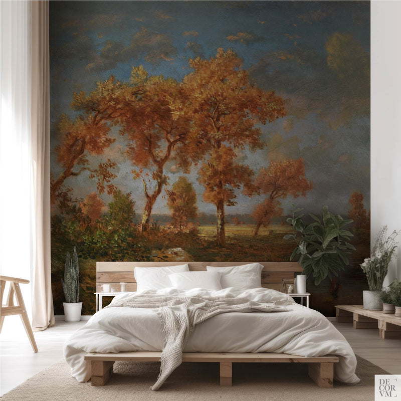 Amber autumn trees wallpaper mural of tall autumn oaks beside a still pond, spanning the wall behind a low timber bed with white linens, a sheer curtain and potted greenery in a soft-neutral bedroom by Decorvm.