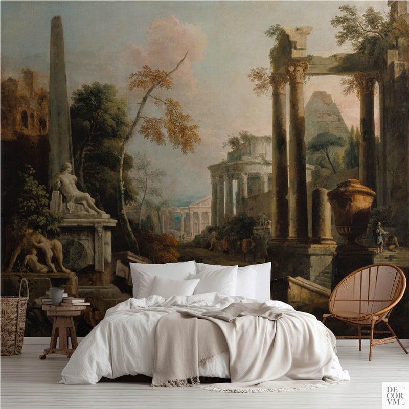 Classical ruins wallpaper mural of Roman columns and a broken obelisk in warm stone, covering the wall behind a low bed with white linens, a wooden stool and a round rattan chair in a soft-neutral bedroom by Decorvm.