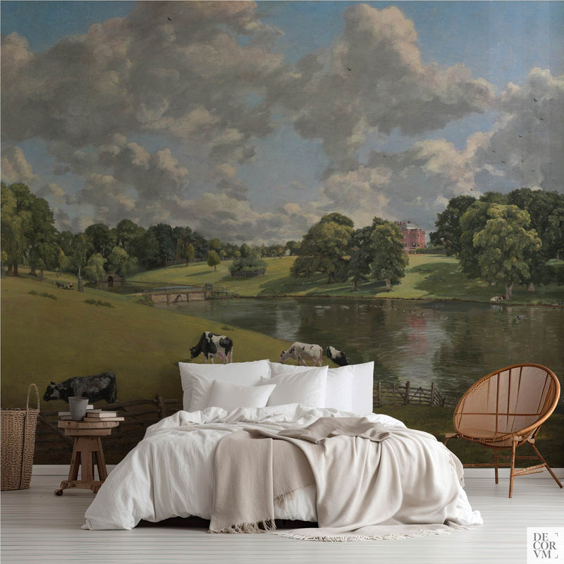 Constable Wivenhoe Park wallpaper mural of an English lake under piled cloud, covering the wall behind a low bed with white linens, a wooden stool and a round rattan chair in a soft-neutral bedroom by Decorvm.