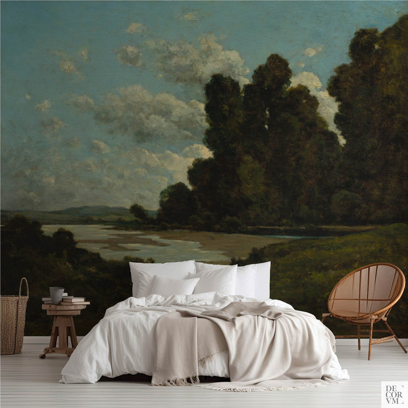 Deep green riverbank wallpaper mural of dark trees over a bright river bend, covering the wall behind a low bed with white linens, a wooden stool and a round rattan chair in a soft-neutral bedroom by Decorvm.