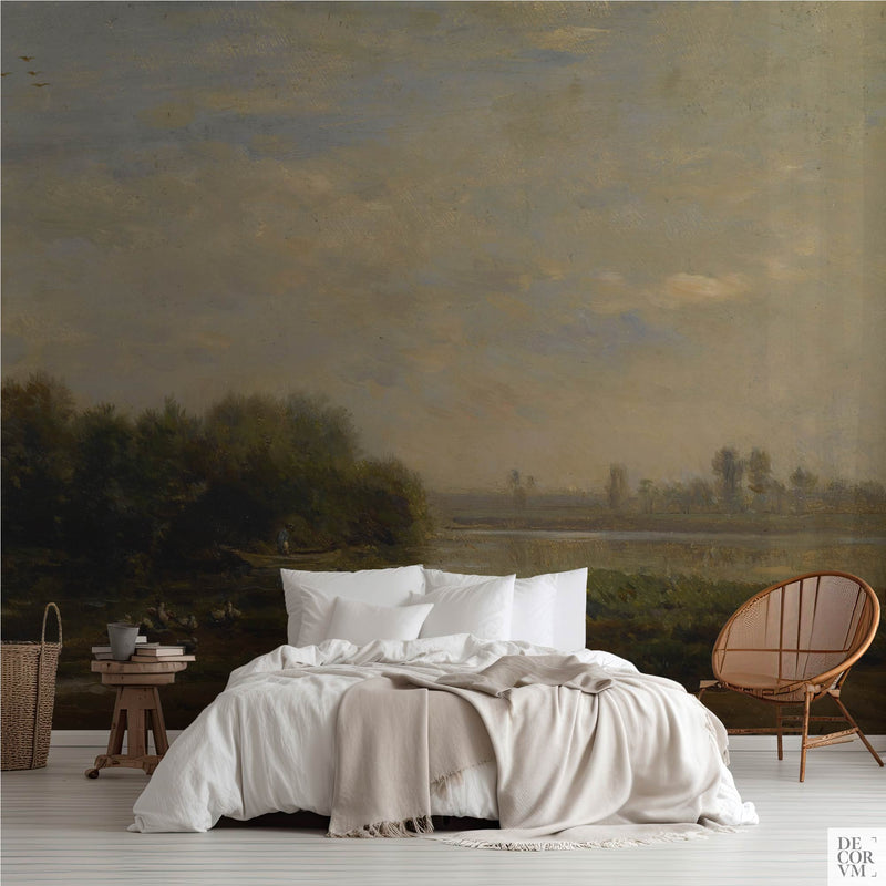 French river ducks pastoral wallpaper mural covering the wall behind a low bed with white linens, a wooden stool, and a rattan chair in a soft-neutral bedroom by Decorvm.