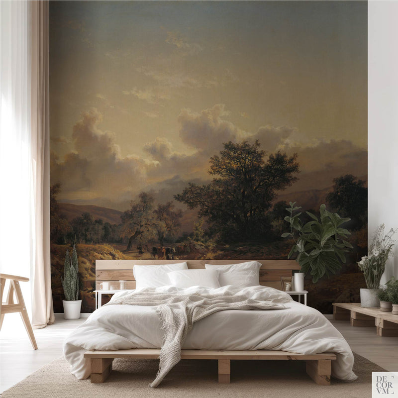 Golden pastoral river wallpaper mural of honeyed countryside and grazing cattle, spanning the wall behind a low timber bed with white linens, a sheer curtain and potted greenery in a soft-neutral bedroom by Decorvm.