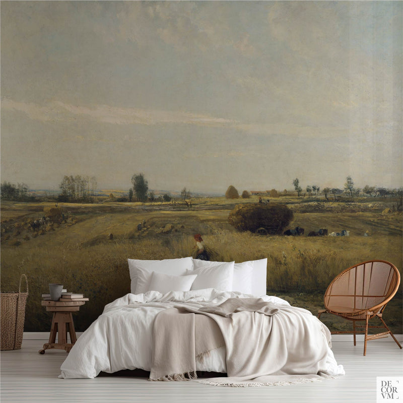 Golden wheatfield harvest wallpaper mural covering the wall behind a low bed with white linens, a wooden stool, and a rattan chair in a soft-neutral bedroom by Decorvm.