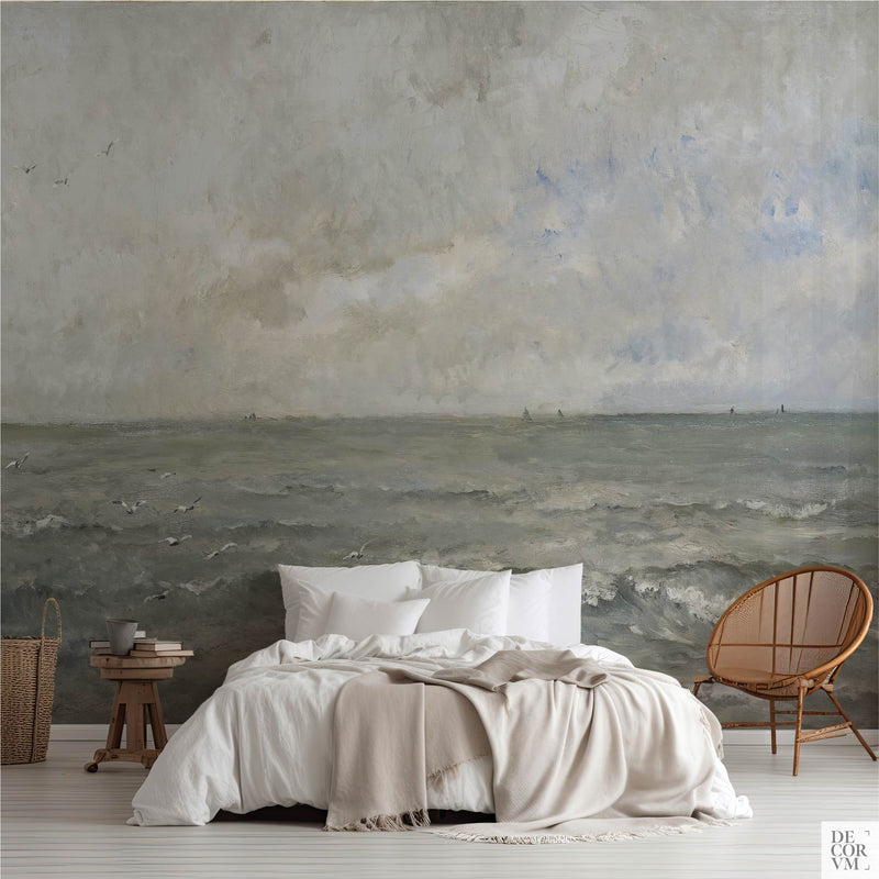 Grey blue sea wallpaper mural of open water under a soft clouded sky, covering the wall behind a low bed with white linens, a wooden stool and a round rattan chair in a soft-neutral bedroom by Decorvm.