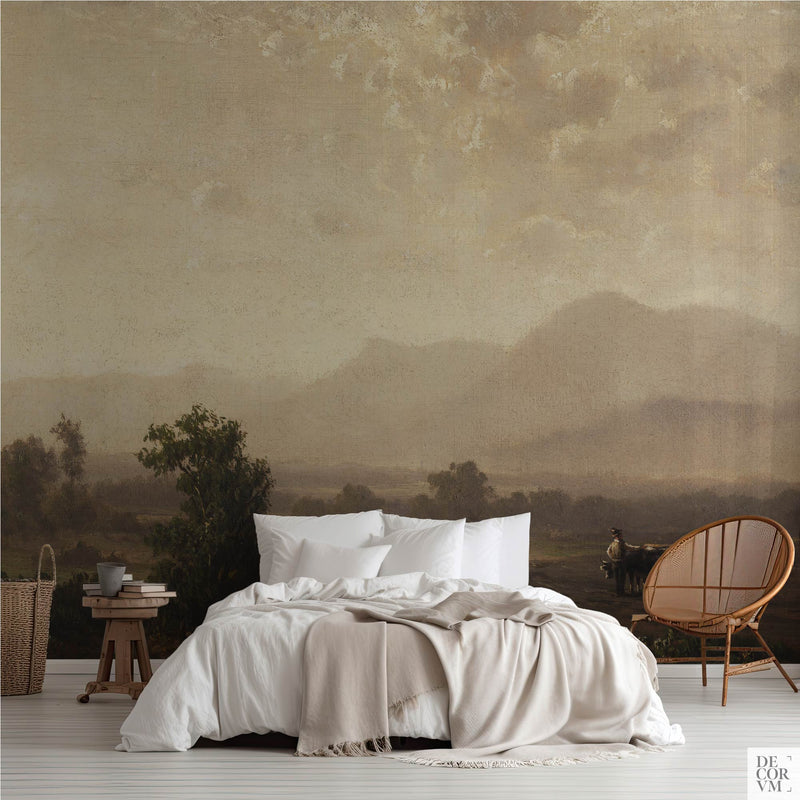 Hazy golden valley wallpaper mural of soft focus hills in warm haze, covering the wall behind a low bed with white linens, a wooden stool and a round rattan chair in a soft-neutral bedroom by Decorvm.