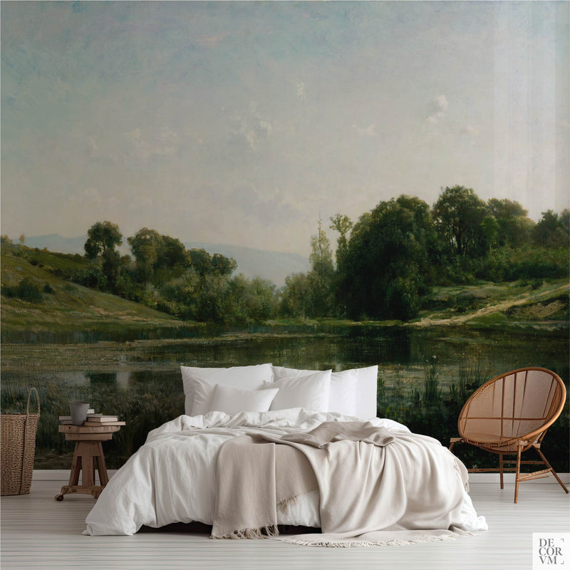 Serene French verdant pond wallpaper mural covering the wall behind a low bed with white linens, a wooden stool, and a rattan chair in a soft-neutral bedroom by Decorvm.
