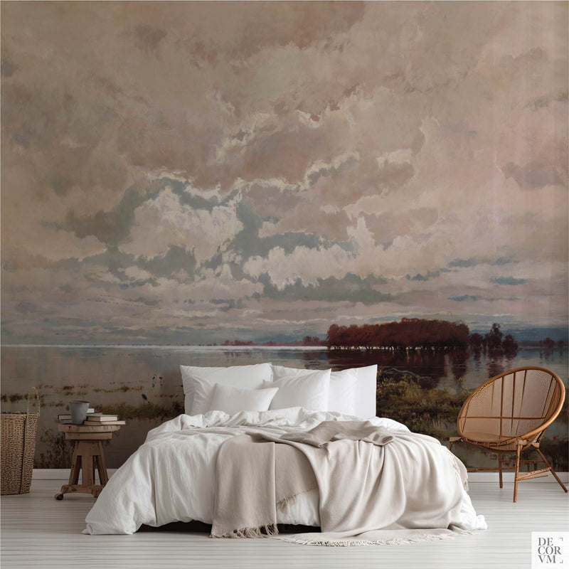 Serene water and sky wallpaper mural of still floodwater under pale cloud, covering the wall behind a low bed with white linens, a wooden stool and a round rattan chair in a soft-neutral bedroom by Decorvm.