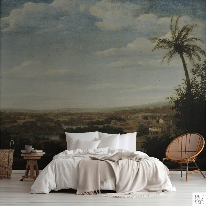 Tropical palm valley wallpaper mural of a tall palm over a wide Brazilian valley, covering the wall behind a low bed with white linens, a wooden stool and a round rattan chair in a soft-neutral bedroom by Decorvm.