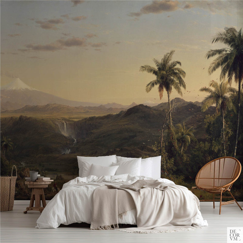 Tropical volcano landscape wallpaper mural of palms and a snow-capped volcano in golden light, covering the wall behind a low bed with white linens, a wooden stool and a round rattan chair in a soft-neutral bedroom by Decorvm.