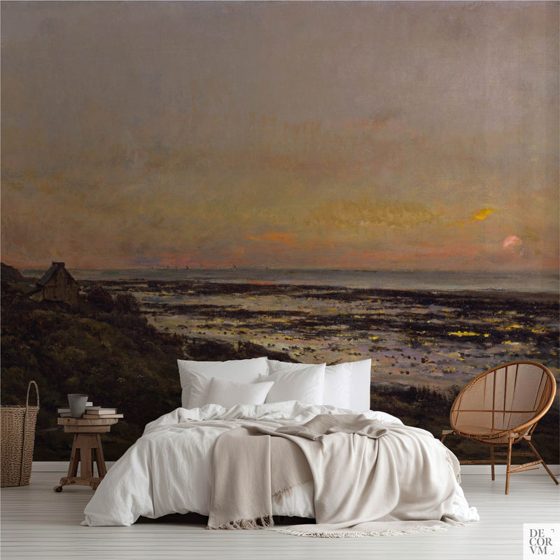 Vintage Normandy beach sunset wallpaper mural covering the wall behind a low bed with white linens, a wooden stool, and a rattan chair in a soft-neutral bedroom by Decorvm.