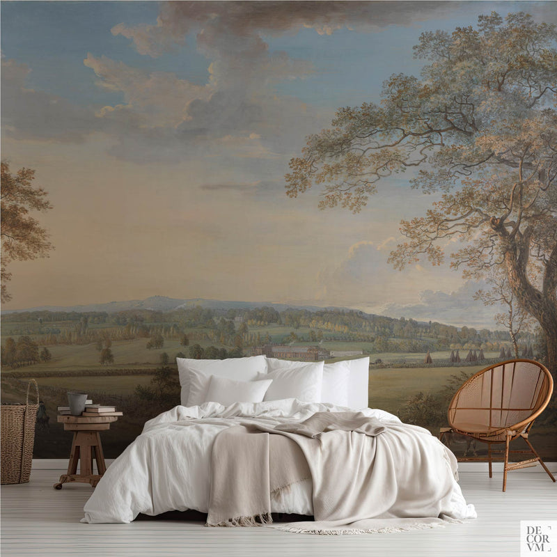 Wide panoramic countryside wallpaper mural of an English parkland vista, covering the wall behind a low bed with white linens, a wooden stool and a round rattan chair in a soft-neutral bedroom by Decorvm.