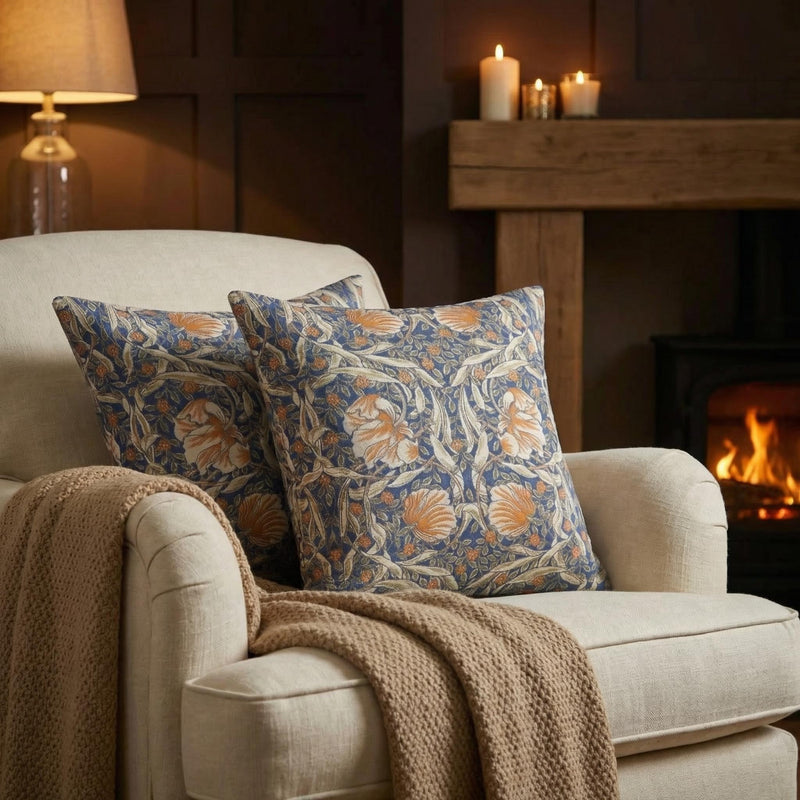 William Morris Pimpernel jacquard throw pillows on a cream armchair beside a fireplace — woven blue and rust 1876 Arts and Crafts pattern by Decorvm.