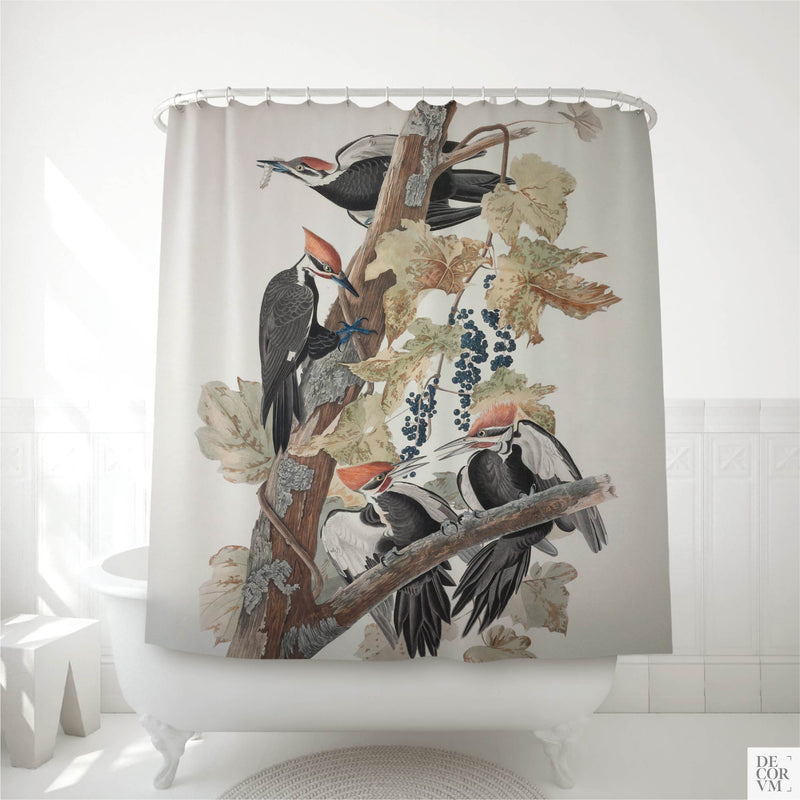 Audubon pileated woodpecker shower curtain displayed in a white bathroom — four woodpeckers scaling a dead tree trunk draped with raccoon grape vine and dark berries, hand-colored engraving on a cream background.