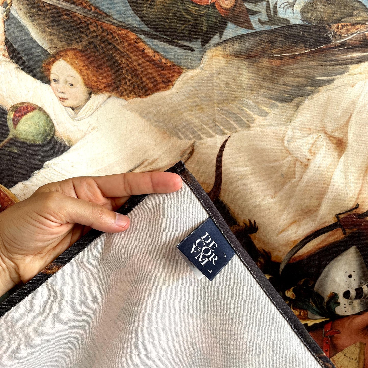 Close-up of a hand lifting the corner of the Bruegel Fall of the Rebel Angels tapestry showing fabric backing and Decorvm woven label.