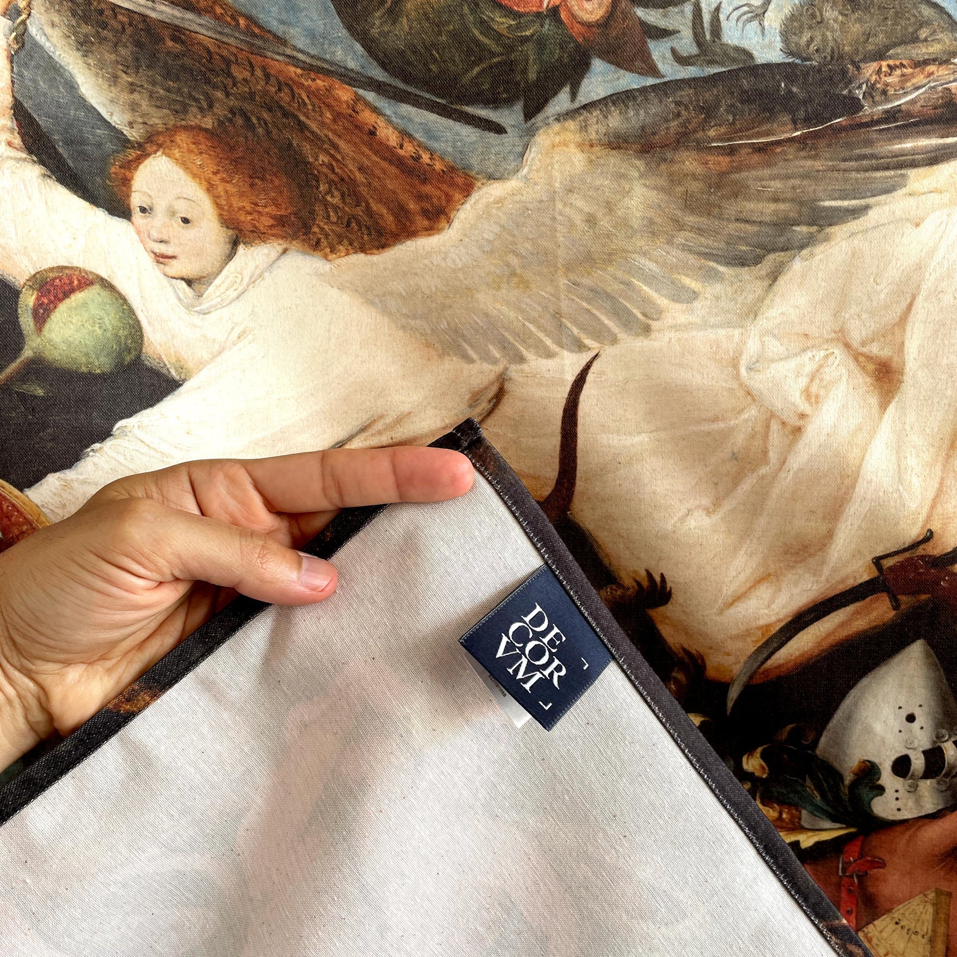 Close-up of a hand lifting the corner of the Bruegel Fall of the Rebel Angels tapestry showing fabric backing and Decorvm woven label.