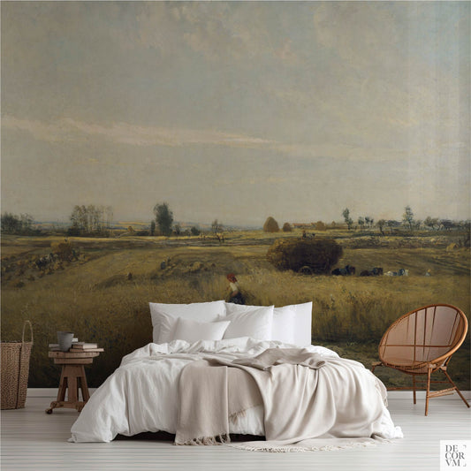 Golden wheatfield harvest wallpaper mural covering the wall behind a low bed with white linens, a wooden stool, and a rattan chair in a soft-neutral bedroom by Decorvm.