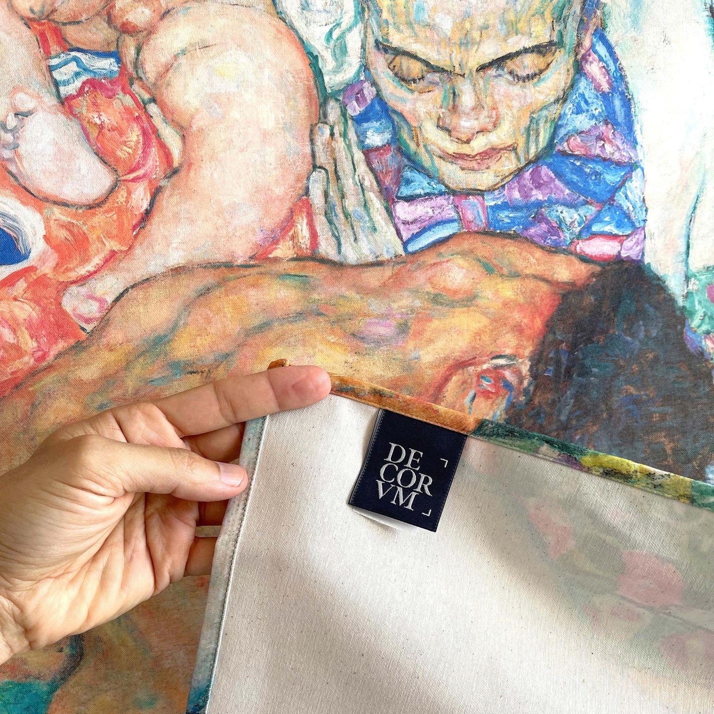 Close-up of a hand holding the edge of the Klimt Death and Life tapestry, showing the printed front, plain white backing, and Decorvm woven label on the hem.