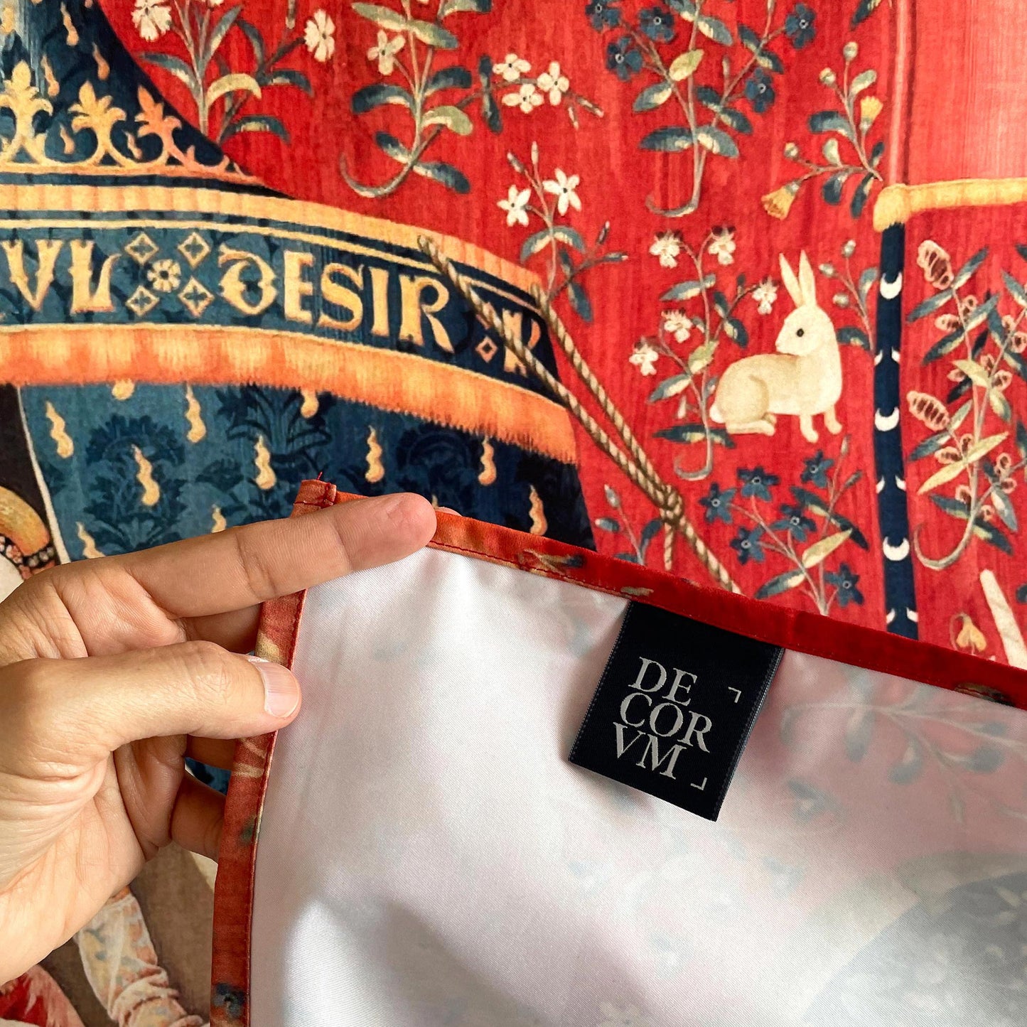Close-up corner detail of the medieval millefleur tapestry showing a hemmed edge, Decorvm brand tag, and a white rabbit on rich red floral background.