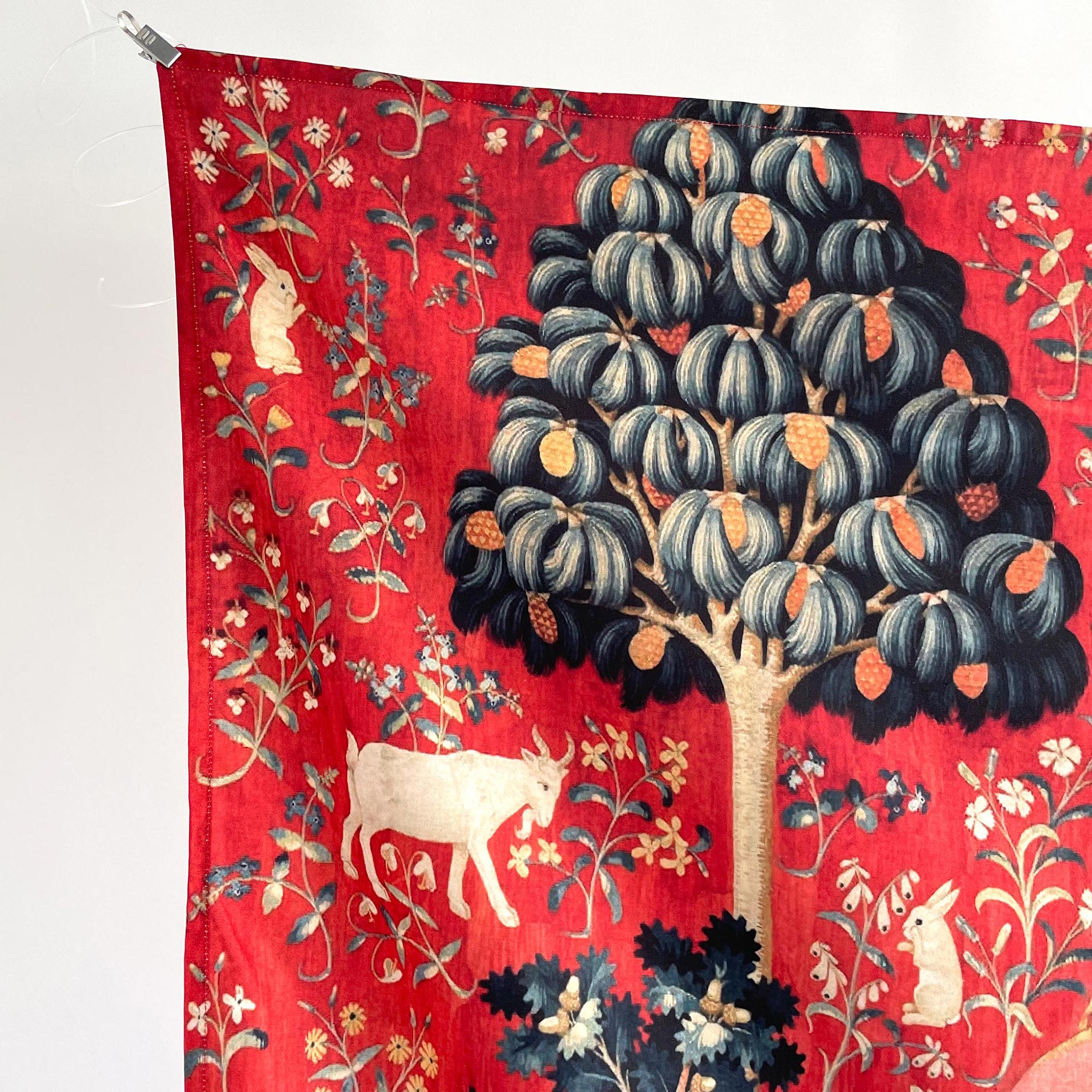 Top-left corner of the red medieval Lady and the Unicorn tapestry — stylized tree with blue foliage, white goat, white rabbit, and millefleur botanicals.
