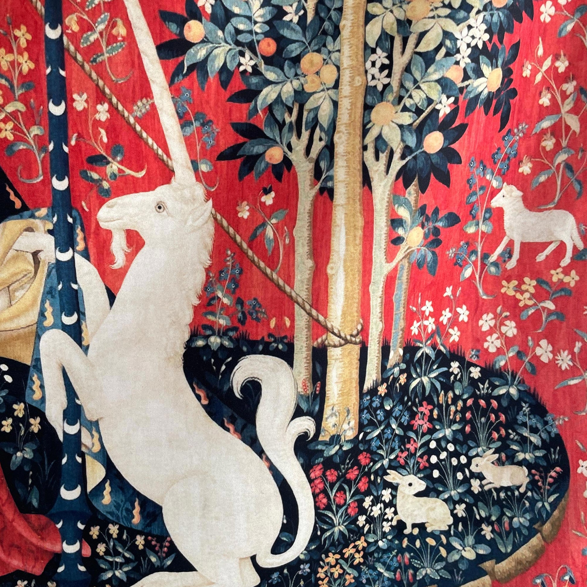 Close-up of the white unicorn rearing up to hold a standard pole on the medieval Lady and the Unicorn tapestry — vibrant red background with traditional medieval flowers and trees.