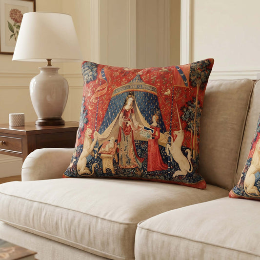 Lady and the Unicorn medieval cushion cover on a beige sofa beside a ceramic lamp — "À mon seul désir" scene with lion, unicorn, and crimson millefleur background.