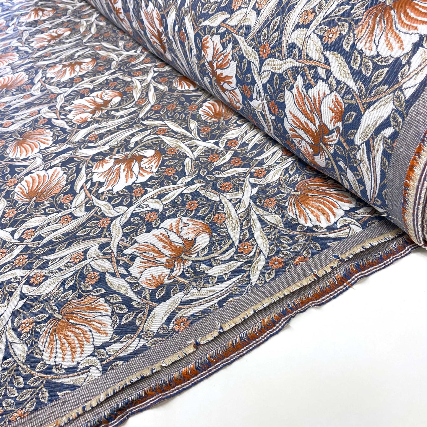 A partially unrolled bolt of Pimpernel Jacquard Fabric displaying a woven floral pattern of rust-colored and white blossoms with swirling leaves on a dark blue background.