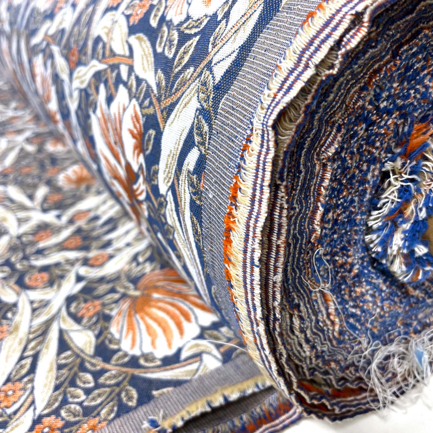 Close-up of the rolled fabric's raw edge, revealing the woven, multi-colored frayed threads of blue, rust, and white that make up the heavy jacquard material.