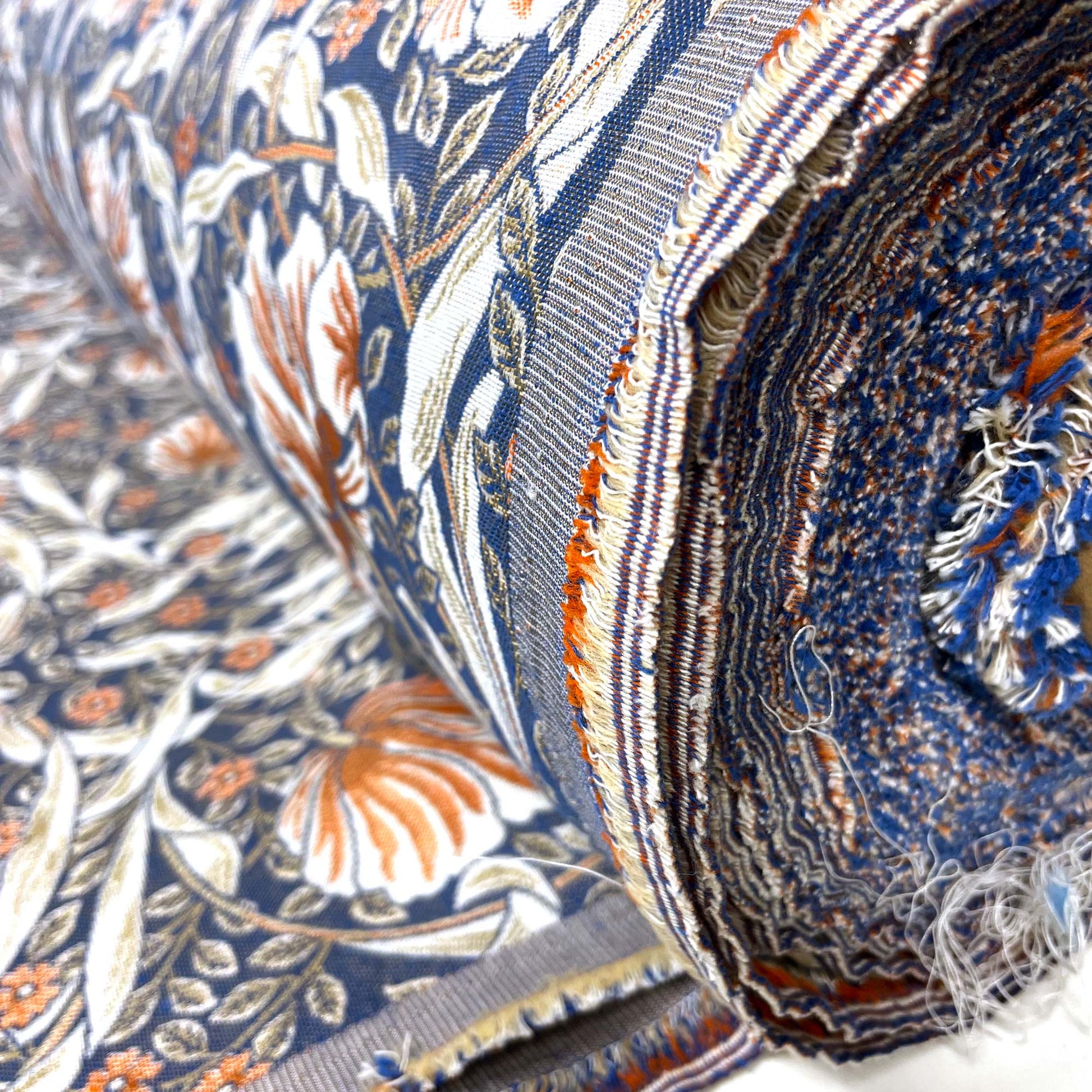 Close-up of the rolled fabric's raw edge, revealing the woven, multi-colored frayed threads of blue, rust, and white that make up the heavy jacquard material.
