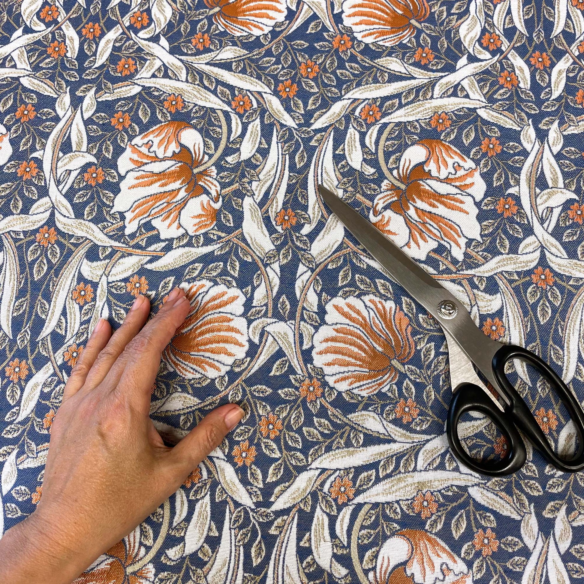 A top-down view of the blue and rust patterned fabric with a person's hand resting on the bottom left and a pair of metal dressmaking scissors laying on the right side.