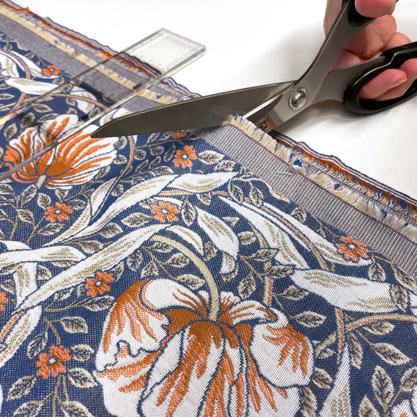A person's hands using metal scissors to cut a straight line across the blue and rust floral jacquard fabric, guided by a clear acrylic sewing ruler.