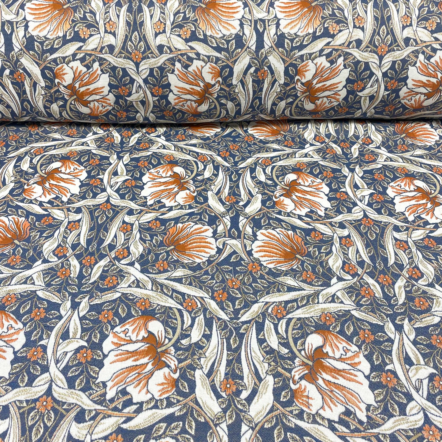 Horizontal view of the Pimpernel Blue Rust jacquard fabric, featuring a rolled bolt at the top unfurling into a flat expanse of the intricate rust-orange and white botanical pattern against a navy blue ground.