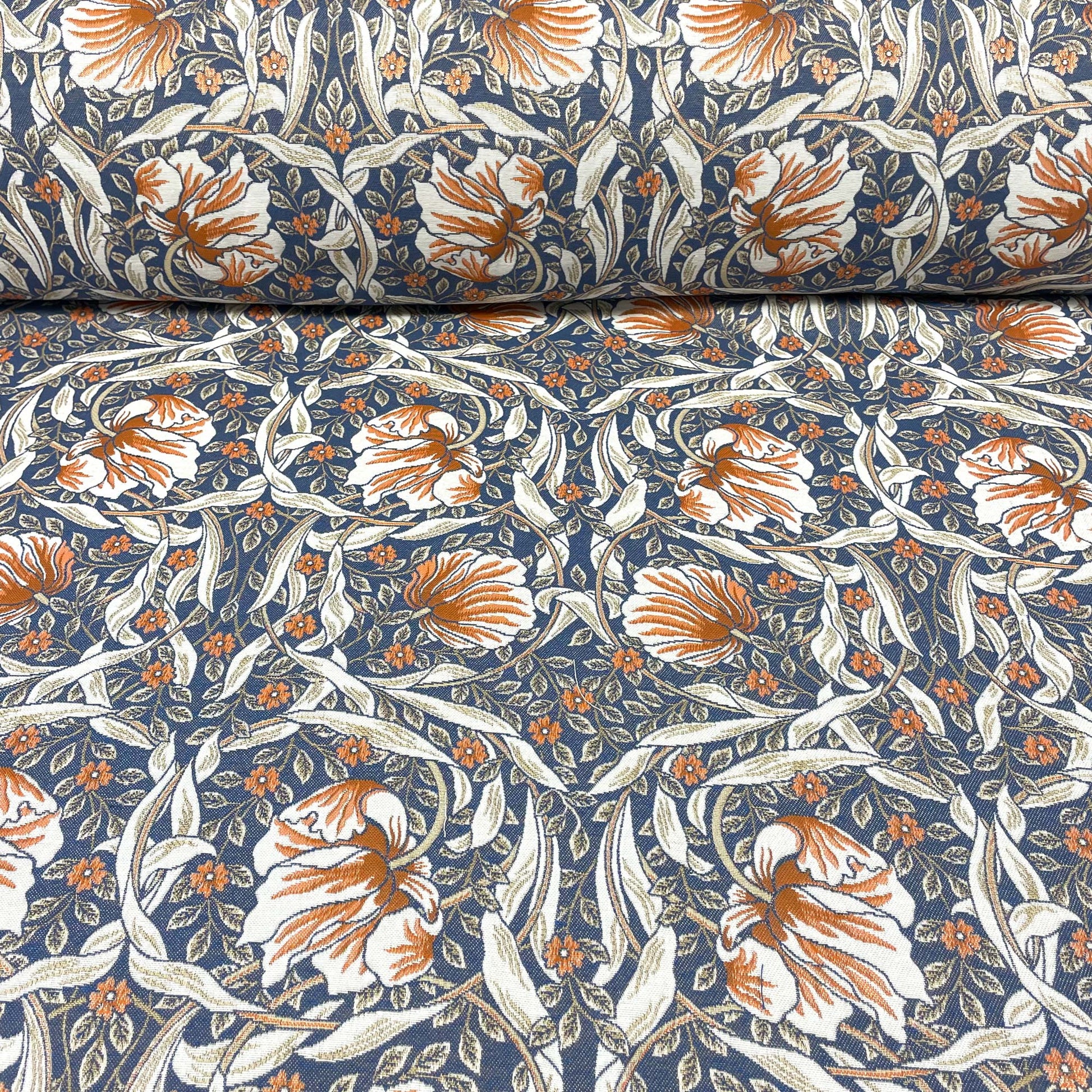 Horizontal view of the Pimpernel Blue Rust jacquard fabric, featuring a rolled bolt at the top unfurling into a flat expanse of the intricate rust-orange and white botanical pattern against a navy blue ground.
