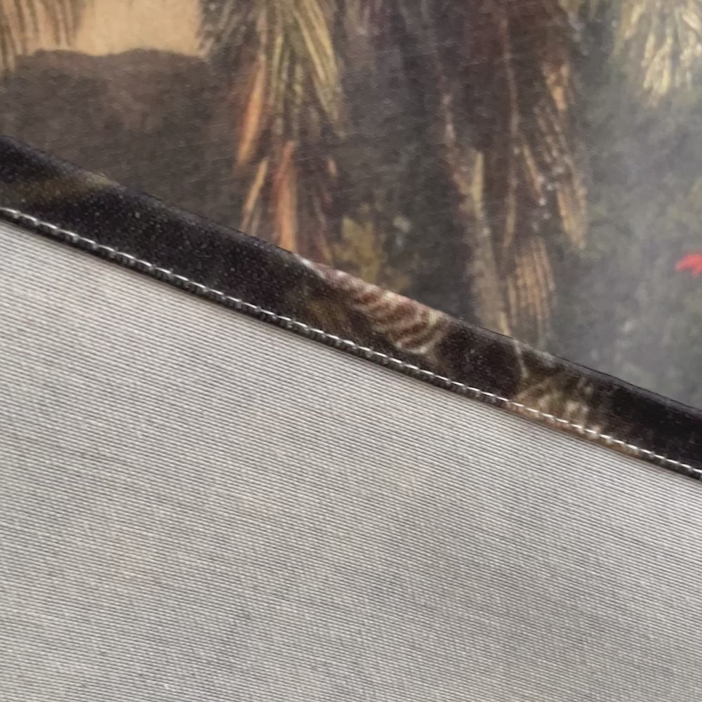 Video close-up panning across tropical volcano landscape wall tapestry showing printed fabric texture, stitched hem detail, palm fronds with exotic birds, single palm tree against misty mountains, snow-capped Cotopaxi volcano with cascading waterfall, and golden amber sky with distant Andean ridgeline