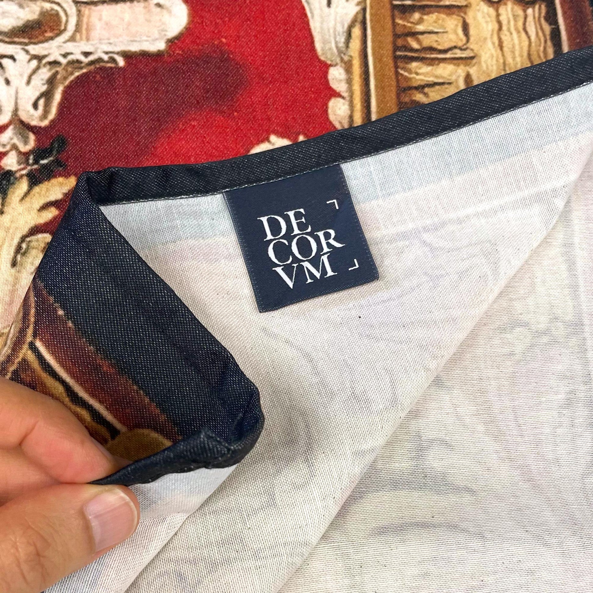 Close-up of the Decorvm brand label on the reverse of The Arms of Louis XV wall tapestry — hemmed dark edge, durable white backing, and a glimpse of the red and gold baroque fabric.