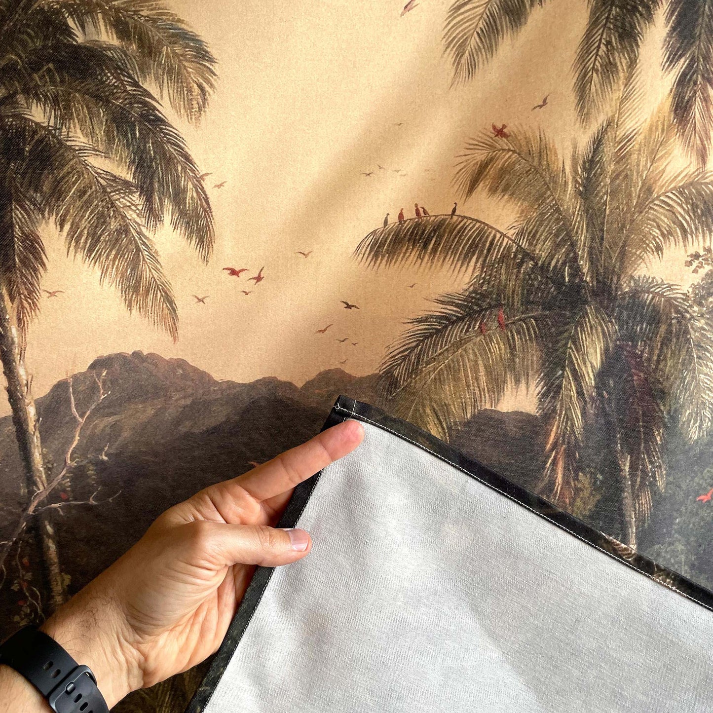 Close-up of palm fronds, flying birds, stitched edge, and reverse fabric of the Frederic Church Cotopaxi wall tapestry, with a hand for scale.