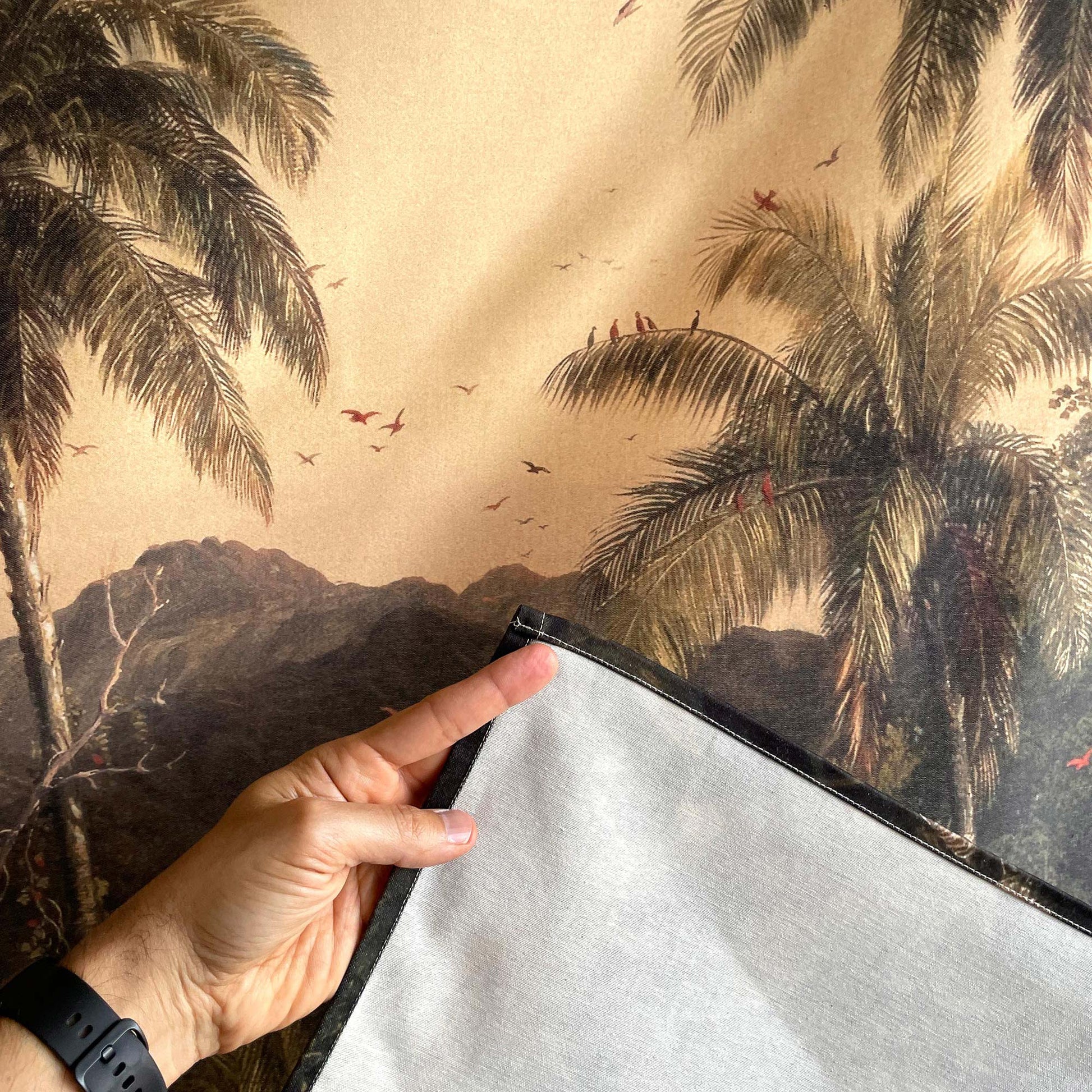 Close-up of palm fronds, flying birds, stitched edge, and reverse fabric of the Frederic Church Cotopaxi wall tapestry, with a hand for scale.