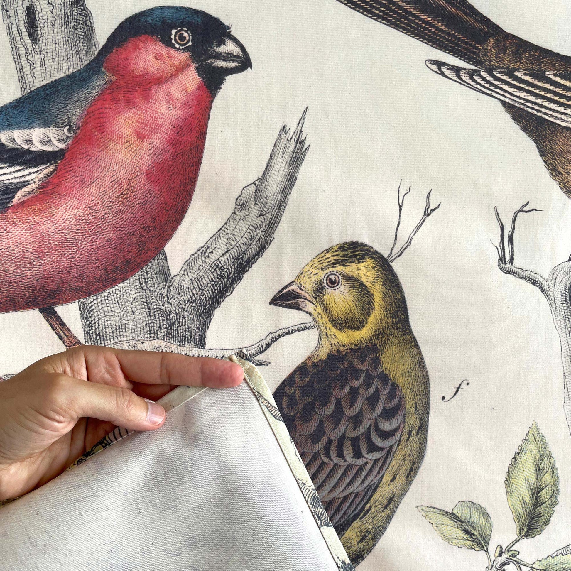 Close-up of the Vintage Ornithology tapestry corner being held back to reveal the unprinted fabric backing, with red and yellow bird illustrations visible on the front.