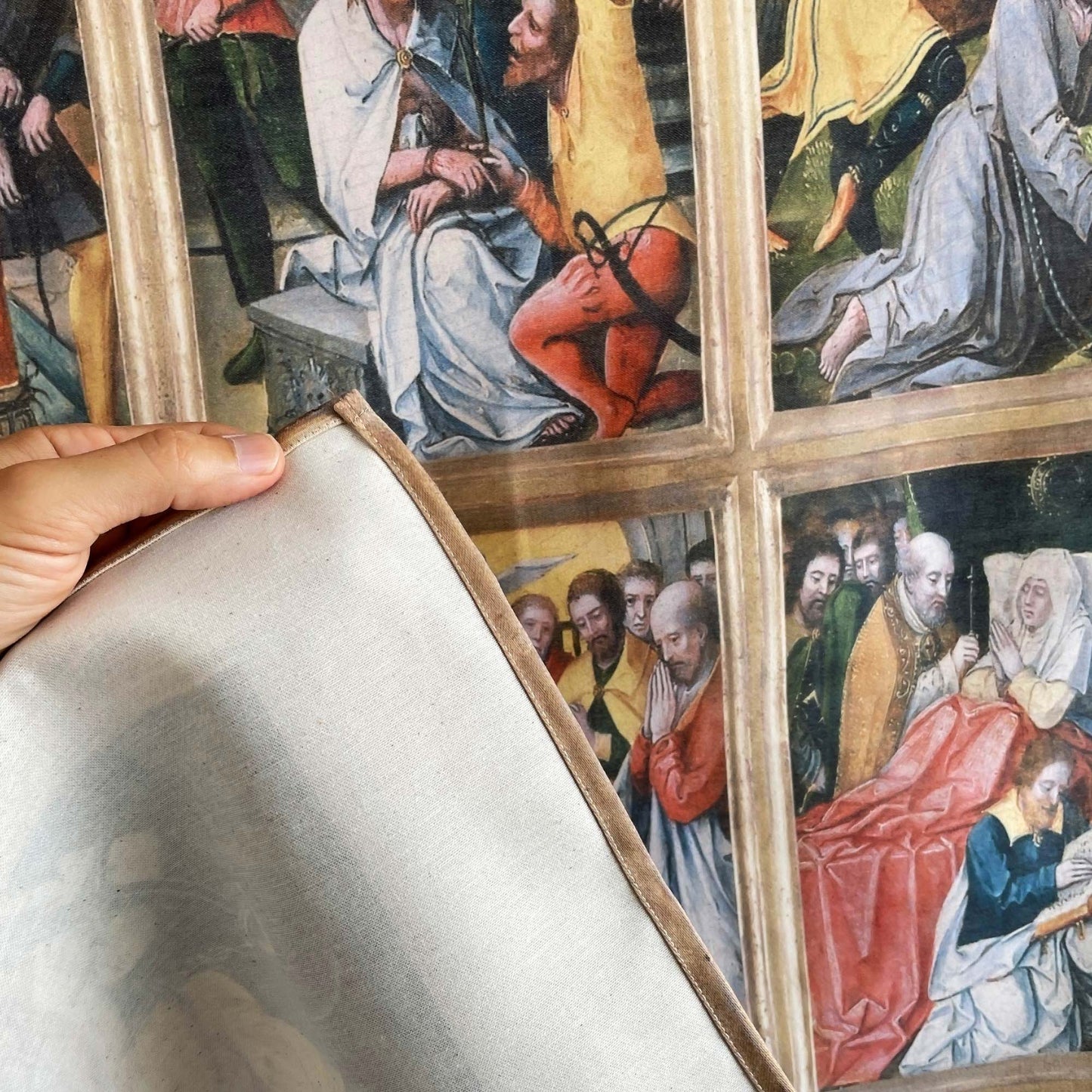 A hand lifting the corner of the Virgin of the Rosary tapestry to show fabric backing and finished edge stitching.