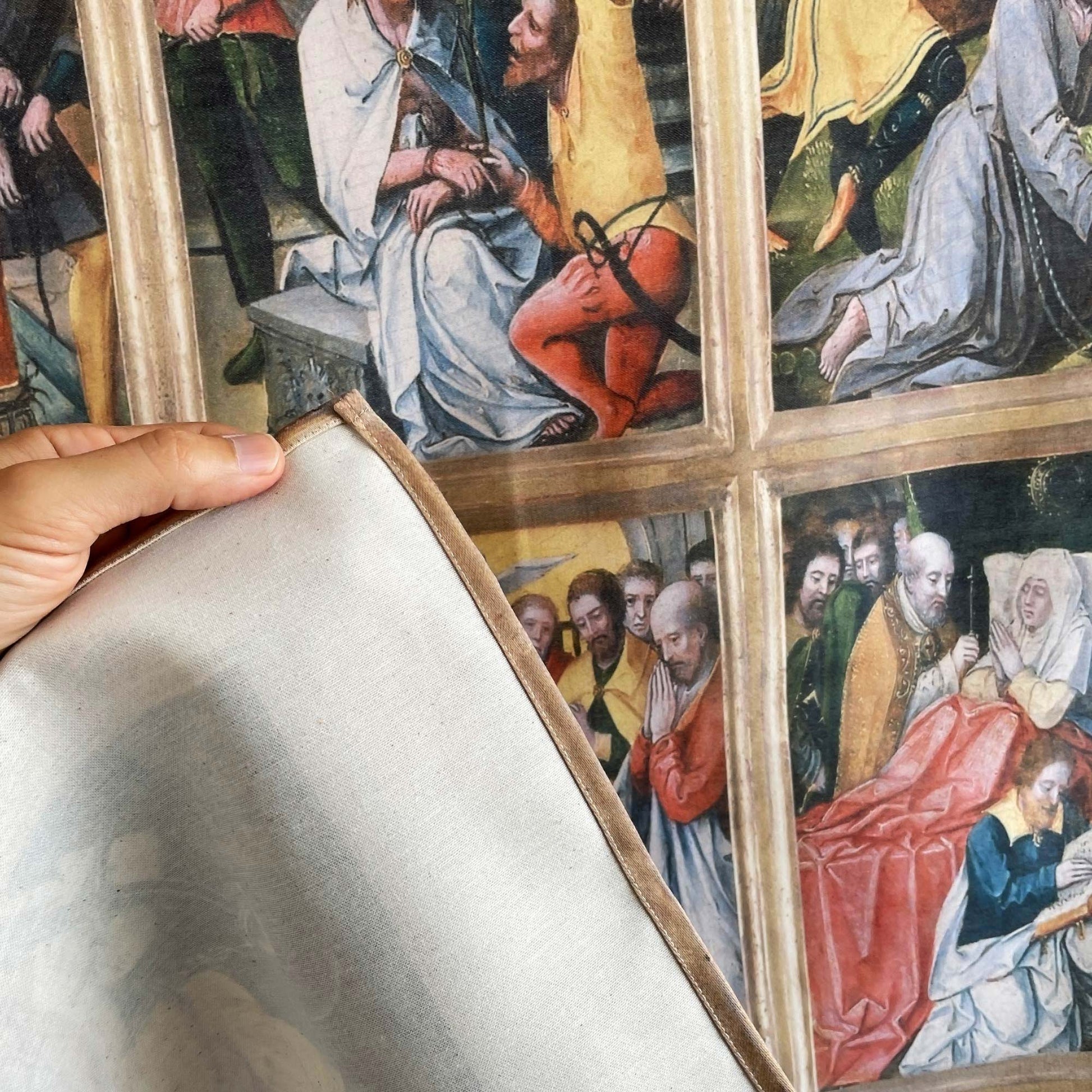 A hand lifting the corner of the Virgin of the Rosary tapestry to show fabric backing and finished edge stitching.