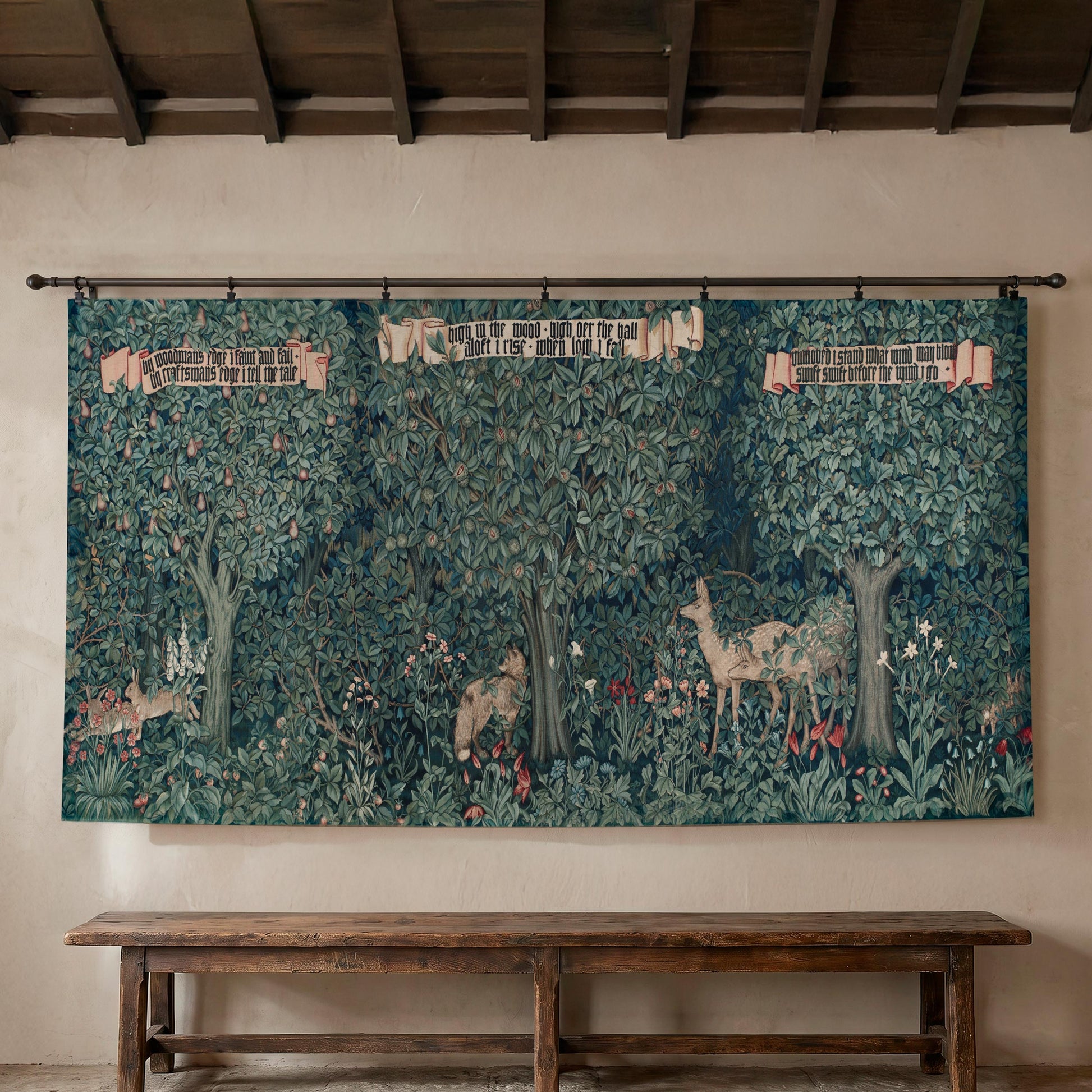 Large William Morris Greenery wall tapestry hung on a textured plaster wall beneath exposed wooden ceiling beams, displayed over a long rustic wooden bench — Arts and Crafts forest with deer, a fox, and botanical detail.